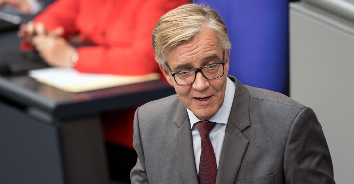 Dietmar Bartsch am Rednerpult des Bundestag, dahinter verschwommen Kanzlerin Merkel in roter Jacke © Soeren Stache/dpa